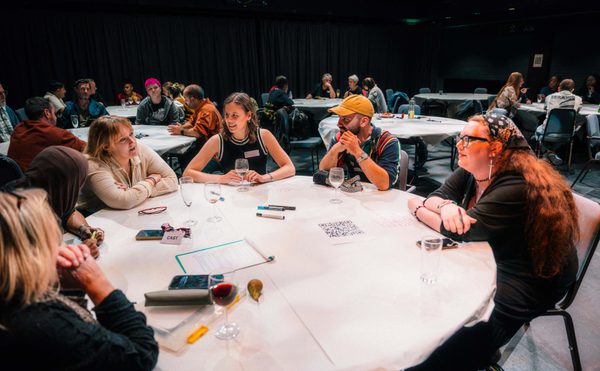 Attendees sit around a large white table at a conference workshop, discussing ideas and reviewing notes in a dim, busy theatre studio.