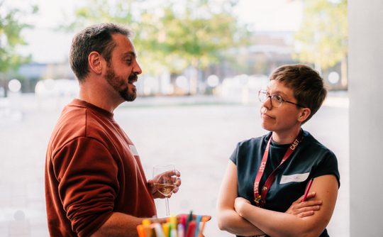 A man in a rust-red jumper chats with a woman in glasses and a dark vest, who listens with arms crossed in a bright, relaxed indoor setting.