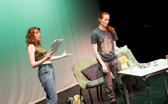 Two women act on stage, holding scripts. One gestures passionately in jeans and a crop top, while the other listens thoughtfully in casual attire.