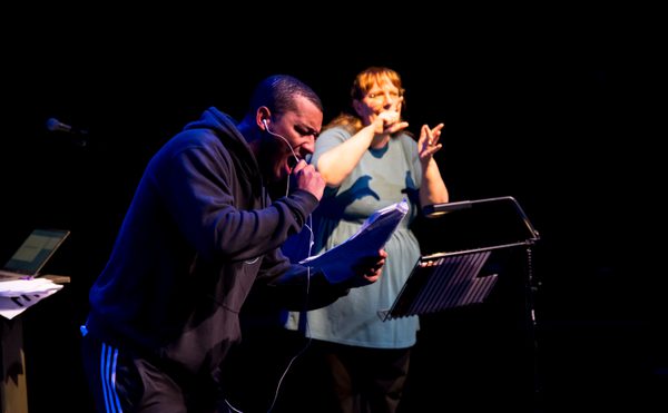 A man in a hoodie passionately speaks into a microphone, while a woman beside him signs BSL energetically on a dimly lit stage, conveying a vibrant performance.