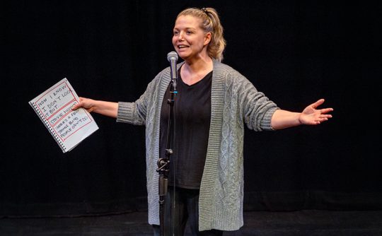 A White female performer in her late 30s wearing a grey cardigan and a blonde ponytail speaks into a microphone. She holds a script.