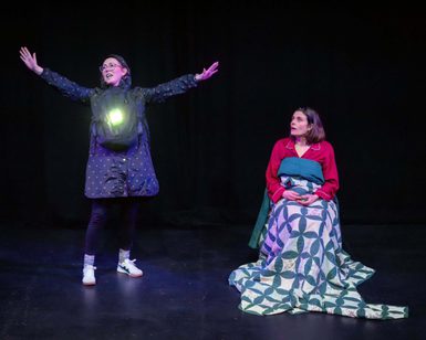 Two White female performers in a black-box studio. One wears all black, standing with arms spread and has a light shining from a bag on her chest. The other woman sits on a chair wearing a red top and wrapped to the chest in a duvet.
