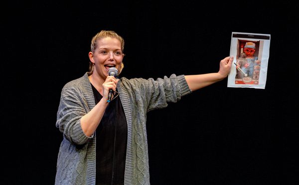 A White woman in her late 30s with blonde hair wearing a grey cardigan. She speaks into a microphone and holds up a picture of a baby doll.