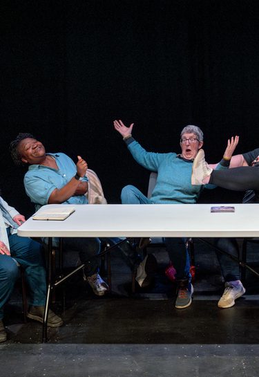 Five older women sit at a long table in front of a black background. The three on the right gesture dramatically. A tartan shopping trolley stands beside the woman on the left.