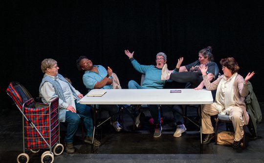 Five older women sit at a long table in front of a black background. The three on the right gesture dramatically. A tartan shopping trolley stands beside the woman on the left.