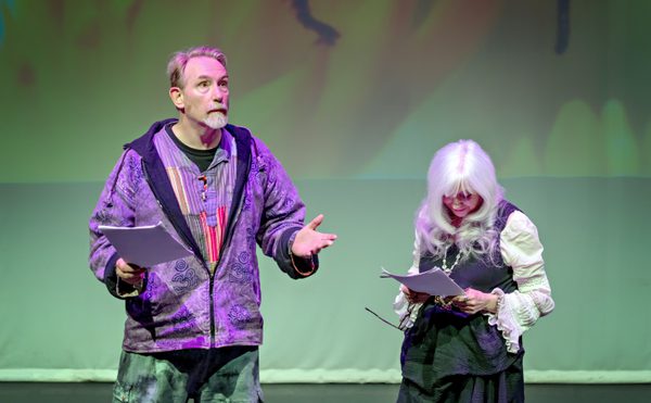 Two people on stage read scripts. The man gestures animatedly, wearing a colourful jacket. The woman, with long white hair, looks focused. The background is softly lit, creating a dramatic atmosphere.