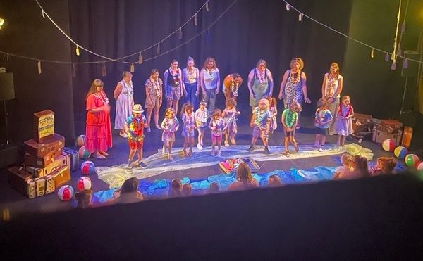 A Cast youth theatre performance featuring young children aged 3 to 5 and their parents on a beach-themed stage with stacked vintage suitcases, beach balls, and hanging string lights.
