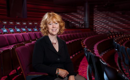 Director Deborah sits in the auditorium surrounded by maroon theatre seating