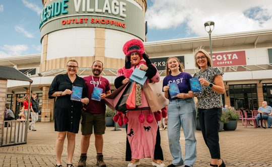 Cast team and staff from Lakeside stand outside Lakeside Village shopping outlet with the panto Dame in a pink dress and wig