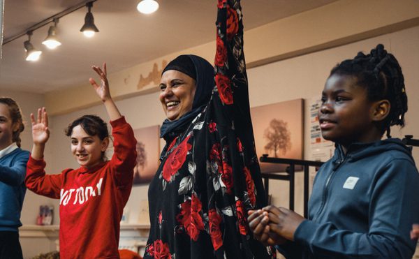 A joyful diverse group of people raise their hands in a room. A smiling woman wearing a hijab and a floral dress stands with children around her, creating an uplifting atmosphere.