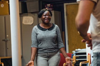 Black woman in gray blouse smiles warmly in a casual room, gesturing with outstretched hands. The atmosphere is welcoming and lighthearted.