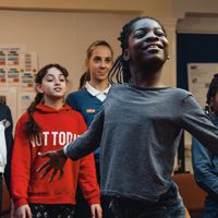 A group of diverse children stand together in a classroom. A smiling black girl in front spreads her arms joyfully, showing confidence and happiness.