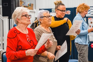 Four women, standing in a row, hold papers and sing in a community setting. They appear focused and engaged, creating a warm and lively atmosphere.