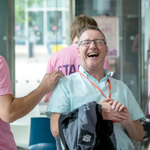 A man in a turquoise shirt laughs joyfully while a person in a pink "STAFF" shirt playfully interacts with him in an indoor setting. The atmosphere is lighthearted.