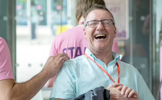 A man in a turquoise shirt laughs joyfully while a person in a pink "STAFF" shirt playfully interacts with him in an indoor setting. The atmosphere is lighthearted.
