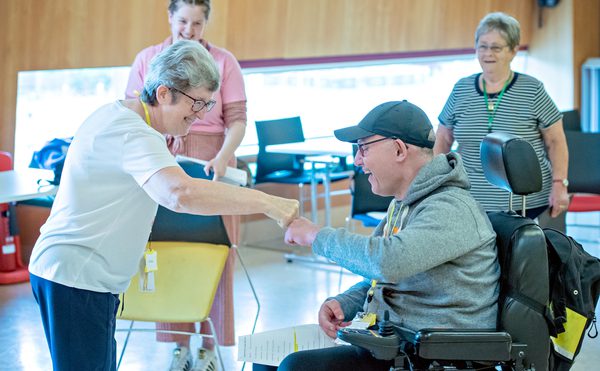 A cheerful interaction in a brightly lit room, where a smiling older woman fist-bumps a man in a wheelchair. Two women observe in the background, creating a warm, inclusive atmosphere.