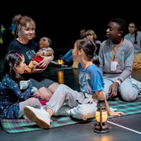 Tech Rehearsals of the Doncastian Chalk Circle. A group of children sit on a checkered blanket, engaged with a woman holding a puppet. A warm, curious atmosphere is conveyed by the soft lantern light.