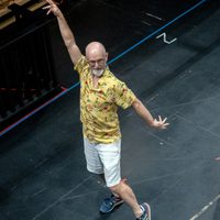Man dancing energetically on stage in a yellow Hawaiian shirt and white shorts, exuding joy and enthusiasm. Background shows stage markings and bags.