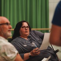 A woman with glasses holds a paper and water bottle, attentively listening, against a green curtain backdrop. Two men are in the foreground.