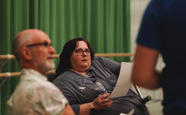 A woman with glasses holds a paper and water bottle, attentively listening, against a green curtain backdrop. Two men are in the foreground.