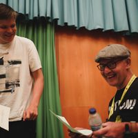 Two people are smiling and holding papers, one is standing the other is sat in a wheelchair there are green and blue curtains behind them. The tone is cheerful and friendly.