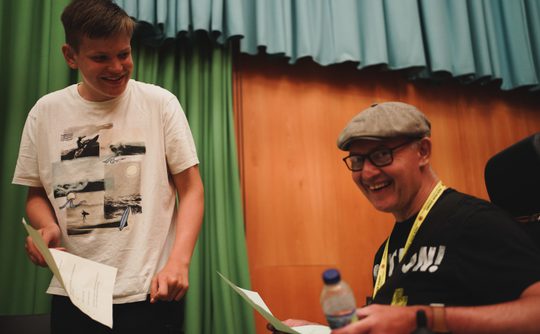 Two people are smiling and holding papers, one is standing the other is sat in a wheelchair there are green and blue curtains behind them. The tone is cheerful and friendly.