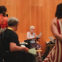A woman with short white hair reads papers in a wood-paneled room, surrounded by others in a discussion, creating a focused, collaborative atmosphere.