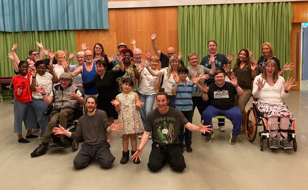 A diverse group of people, some in wheelchairs and some seated and standing, smiling and raising their hands in a joyful pose. The background features curtains in blue and green tones at the front are two members of the Jasmin Vardimon company whoi lead a workshop with the group.