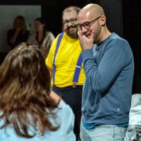 Two men smiling and laughing, one in a blue sweater, the other in a yellow shirt and purple suspenders, enjoying a casual, joyful moment indoors.