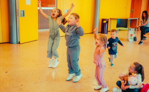 Five young children joyfully jump and cheer in a bright room with yellow walls. A seated woman in the background smiles, creating a lively atmosphere.