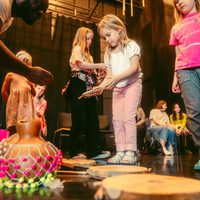 Children engaged in a group drumming activity lead by a black man in colour clothing, surrounded by adults. A decorative gourd with beads is on the floor, creating a playful atmosphere.