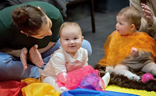 A smiling baby sits on multicoloured fabric, playing with purple gauze. There is a clapping parent to the left and another baby in orange gauze to the right.
