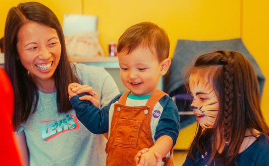 A grinning mother gazes at a smiling toddler in orange dungarees. A girl to the right with tiger face paint helps the toddler bang a drum.