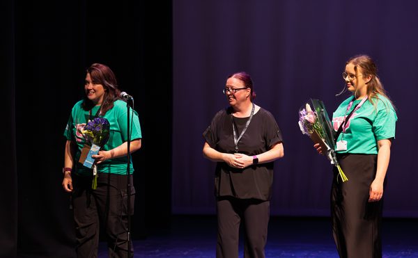 Three women on stage during an event. Two hold bouquets and wear green "Speak Up" t-shirts; the third is in black, smiling. The atmosphere is joyful and celebratory.