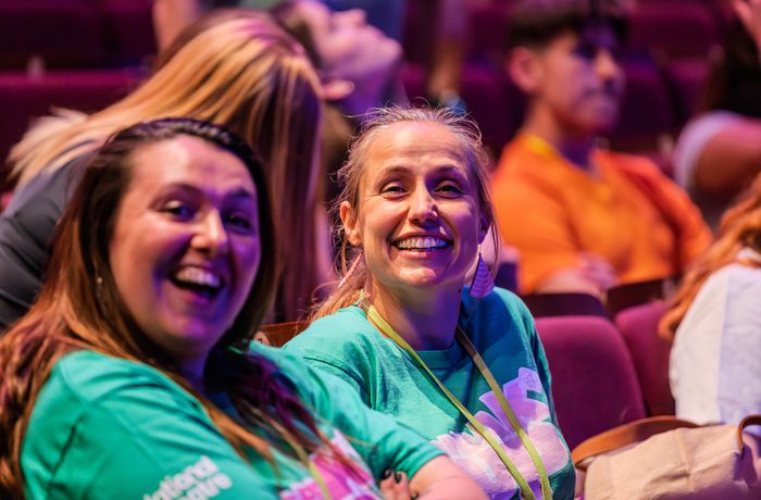 Two women in green "Speak Up" shirts sit smiling on purple theater seats, exuding joy and excitement. A blurred crowd in the background creates a lively ambiance.