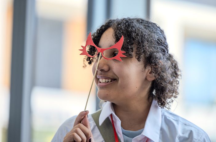 Young person holding red novelty glasses on a stick, wearing a white shirt, with curly hair, indoors with blurred windows behind them. Playful mood.