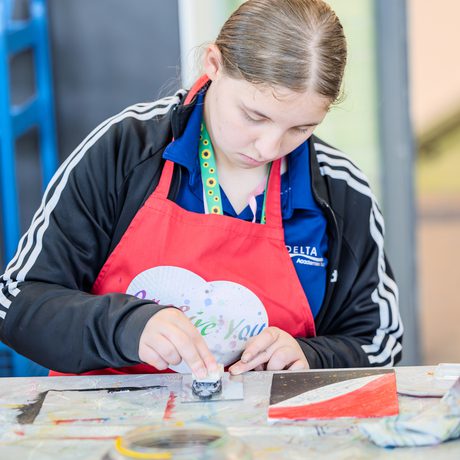 A focused young person in a red apron works on an art project, painting carefully on a table. The setting is bright and creative, conveying concentration.
