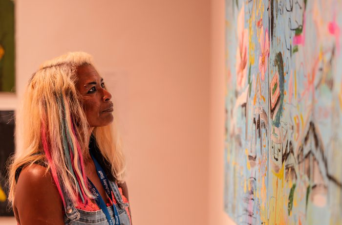A person with colorful hair intently observes a vibrant abstract painting in an art gallery. The scene conveys curiosity and appreciation for art.
