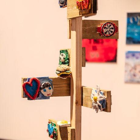 Wooden post with colorful clay art pieces attached, including a heart, face, and flower. Art on display at Doncaster's Gallery and Museum, warm lighting, creative and lively atmosphere..