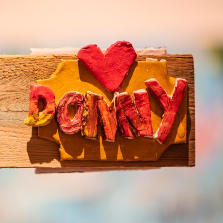 Wooden sign displaying a sculpture that says "Donny" in textured clay, red letters beneath a red heart, set on a sunny, warm-toned background, conveying a feeling of affection for home and Doncaster..