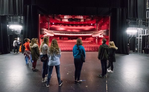A group of people stand on a dimly lit stage, facing an empty theater with red seats. The theater's warm glow creates a contemplative atmosphere as they tour the building.