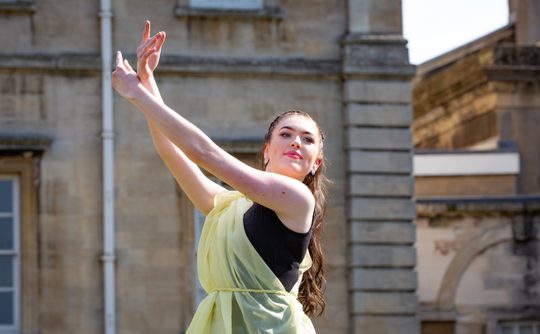 A female dancer in a flowing yellow dress poses gracefully, arms elegantly lifted against a backdrop of historic stone architecture. The scene conveys elegance and poise.