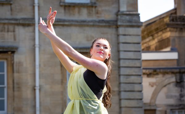 A female dancer in a flowing yellow dress poses gracefully, arms elegantly lifted against a backdrop of historic stone architecture. The scene conveys elegance and poise.