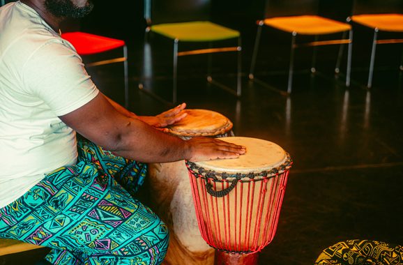 A black man in colorful geometric pants plays two African drums with focus. Brightly colored chairs are in the background, suggesting a lively atmosphere.