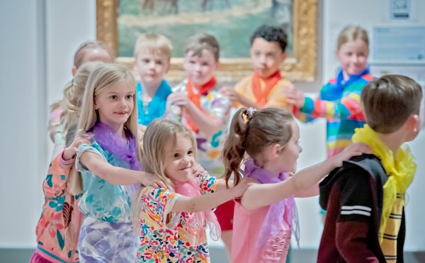 A group of children walk in a circle with their hands on each others shoulders. They smile excitedly and they perform to the audience. Behind them is a large wall with a beautiful painting displayed. This photo is from the 2023 Cast Youth Theatre Production of Time Casters performed at the Danum Gallery Library and Museum.