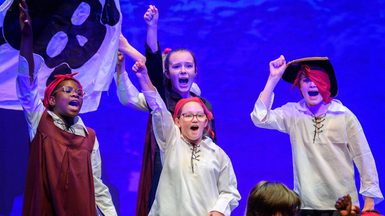 Children dressed as pirates perform on stage, exuberantly raising fists against a purple backdrop with a skull flag, conveying excitement and adventure. Photo is from the 2023 Youth Theatre production of Time Casters.