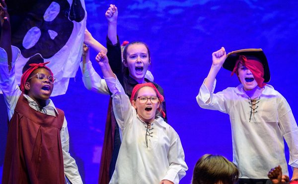 Children dressed as pirates perform on stage, exuberantly raising fists against a purple backdrop with a skull flag, conveying excitement and adventure. Photo is from the 2023 Youth Theatre production of Time Casters.