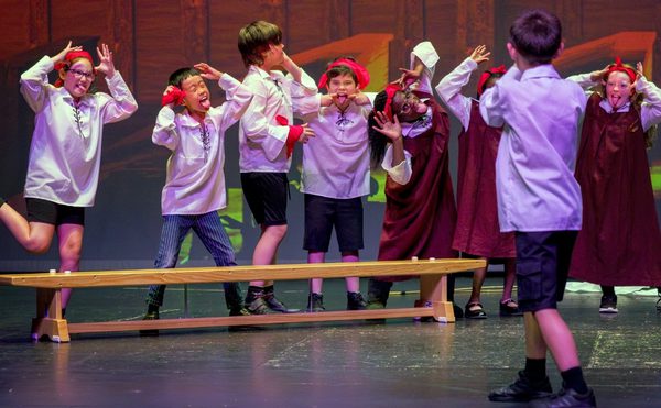 Children in red headbands and white shirts with red gloves perform a playful and energetic scene on stage, making silly faces and gestures. A lively and humorous atmosphere is conveyed. Photo is from the Youth Theatre 2023 production of Time casters.