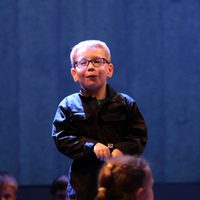 Young child with glasses on stage wearing dark clothing, looking thoughtful. Dim lighting and blue background create a focused, dramatic mood. This photo is from the 2019 Cast Youth Theatre Production of We Are Giants.
