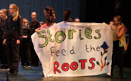 A group of children on stage, some holding a banner reading "Stories Feed the Roots" with colorful lettering and a flower design, evoking creativity. This photo is from the 2019 Cast Youth Theatre production of We Are Giants.